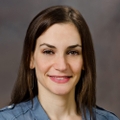 Shana Kusin has long brown hair and smiling against a brown background.