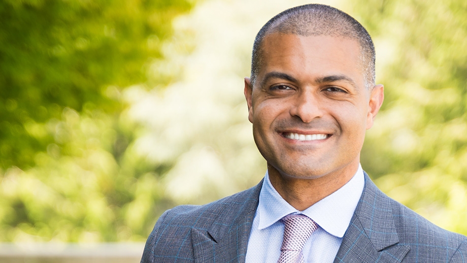 Shereef Elnahal, M.D., M.B.A., tiene el cabello oscuro afeitado, viste un traje gris claro con una camisa azul claro y corbata y sonríe en un área de jardín en OHSU.