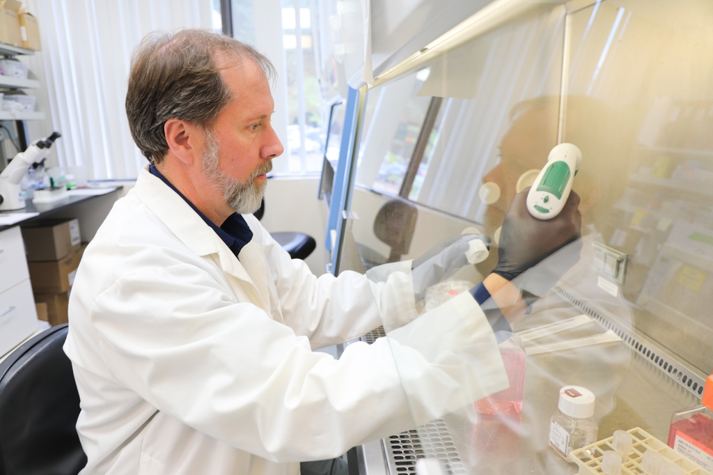 Mark Slifka, Ph.D., tiene cabello castaño corto y fino, vello facial castaño y blanco, viste una bata blanca y realiza algunas tareas de pipeteo bajo una capucha en su laboratorio.