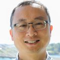 Zheng Xia, Ph.D., tiene cabello oscuro muy corto, anteojos, una camisa azul y verde y sonríe con el Puente Tillicum detrás de él.