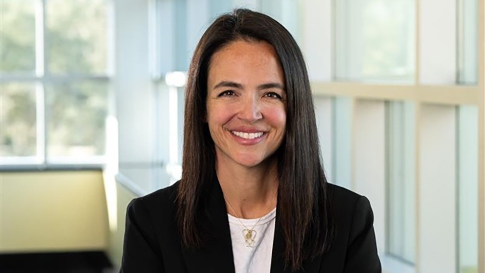 Angela Sklenka has long, straight, brown hair, a white top and black blazer, smiling near a window.