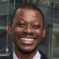 Lishomwa Ndhlovu, M.D., Ph.D., has on a white shirt, black pants and blazer, smiling with arms folded in a stairway