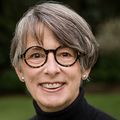 Ann has short gray hair, eye glasses and a black sweater, smiling in a garden.