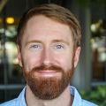 James Frank, Ph.D. has short red hair and facial hair, a blue shirt and smiling in a garden.