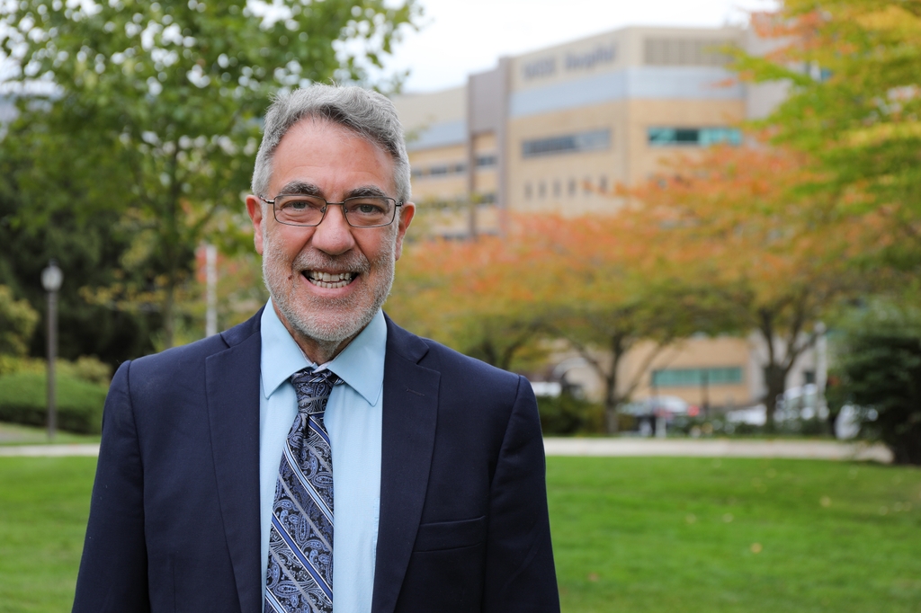 William Hersh, M.D., has gray hair and facial hair stubs, eye glasses, and a blue suit and tie with a light blue shirt, smiling on the grass with OHSU behind him.