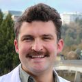 Samuel Melville tiene el pelo corto, rizado y castaño, lleva bigote y viste una bata blanca.