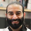 Karimi tiene el pelo largo recogido, lleva una bata de laboratorio y está sentado sonriendo en su laboratorio.