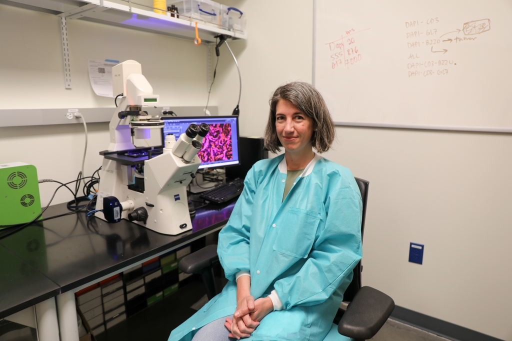 Katelyn has cropped light brown hair, a turquoise lab gown and is sitting at a microscope.