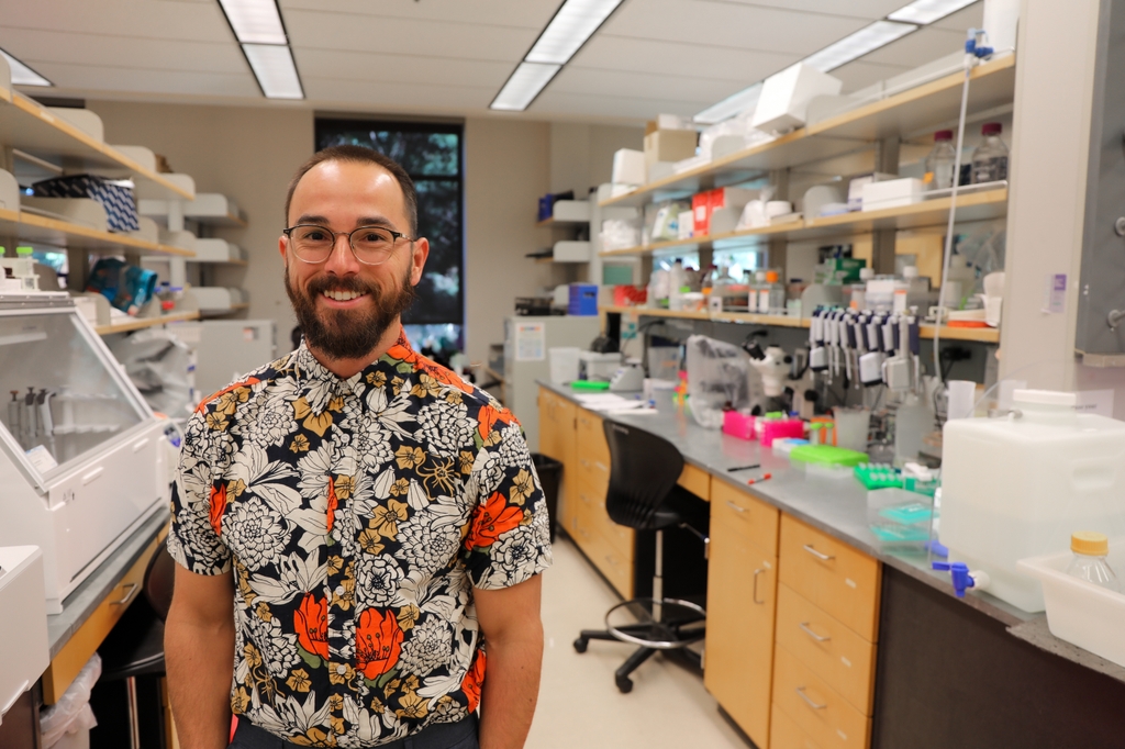 Brandon Wilder, Ph.D., an immunologist and associate professor in OHSU's Vaccine and Gene Therapy Institute, studies malaria with the goal of developing an effective vaccine. (OHSU/Christine Torres Hicks) Brandon has short brown hair, facial hair, eye glasses and a black/white/orange print shirt. He is smiling in his laboratory.