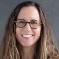 Barbara has long light brown hair, eye glasses, and is wearing her white coat with a black shirt under.