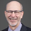 Kyle has a bald hairstyle, gray facial hair, eye glasses and is wearing a dark blue suit and tie.