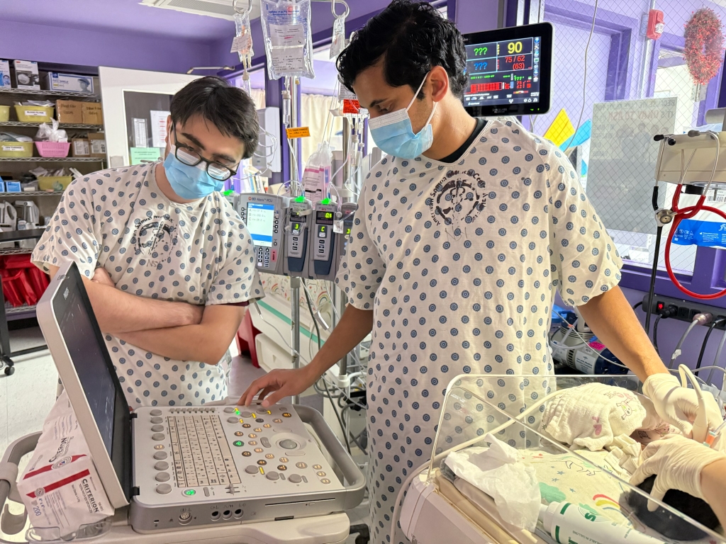 Austin, a la derecha, observa el equipo en una UCIN, de pie junto a la cama de un paciente infantil.
