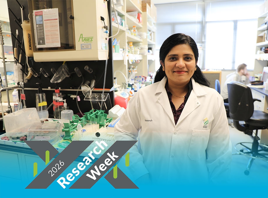 Meghna has long dark hair pulled back away from her face, and is smiling in her lab, wearing a white coat.