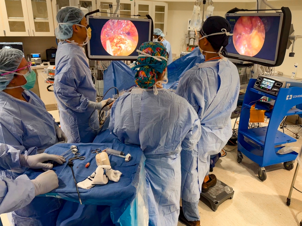 A group of surgeons in blude caps and scrubs gather around a patient in a surgery room at OHSU, looking at image monitors during surgery.