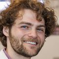 Gabriel tiene el pelo castaño, espeso y rizado, y lleva una bata blanca mientras sonríe a una mesa de laboratorio en OHSU.