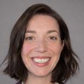 Anna has short dark hair, cropped and is wearing a brown blazer and cream top, smiling.