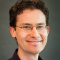 Brian tiene el pelo corto, castaño y rizado, usa gafas y lleva traje.