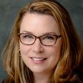 Jennifer has long, curly red-blonde hair, eye glasses, and a black suit with brown top, smiling in a photo studio.