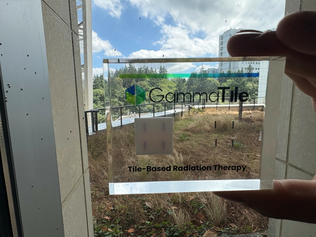 Una persona sostiene un GammaTile dentro de una funda transparente, cerca de una ventana. Los edificios y árboles de OHSU se encuentran al fondo.