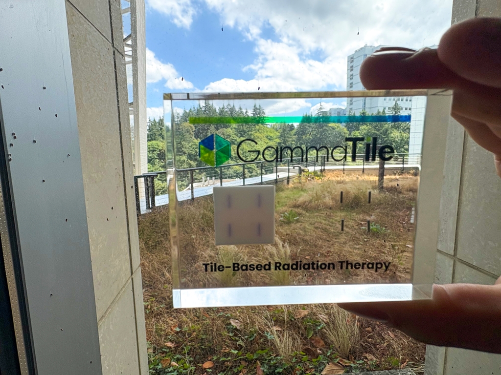 A person holds up a GammaTile device that is in a clear case, near a window. OHSU buildings and trees in the background. subject-centered