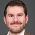 Joe has short brown hair, a brown suit jacket, a white dress shirt and a bolo tie. He is smiling in a photo studio that has a gray/brown background.