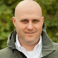 Joe has no hair, a white dress shirt and green jacket, smiling in a garden.