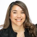 Angelica has long brown hair, a white shirt and black jacket, smiling against a white background.