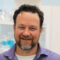 Trevor McGill, Ph.D. (OHSU) McGill has short wavy brown hair, brown/gray facial hair, and is wearing a blue dress shirt, smiling in his lab.