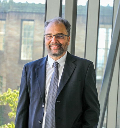Fabrice tiene el cabello corto y canoso, vello facial y gafas; viste un traje gris con camisa blanca y corbata gris azulada, y sonríe mientras se encuentra en la pasarela interior que conecta el Sam Jackson Hall con el Lamfrom Biomedical Research Center.