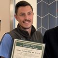 Tyler has short brown hair and a green nursing uniform, standing and holding his certificate, surrounded by colleagues. 