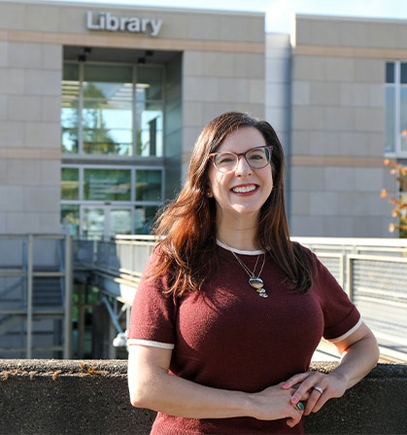 Kristin tiene el cabello largo de color marrón oscuro, lleva gafas, una blusa color burdeos y pantalones marrones, y sonríe frente al edificio de la Biblioteca de OHSU.