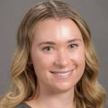 Claire has wavy blonde hair and a brown shirt, smiling in a photo studio.