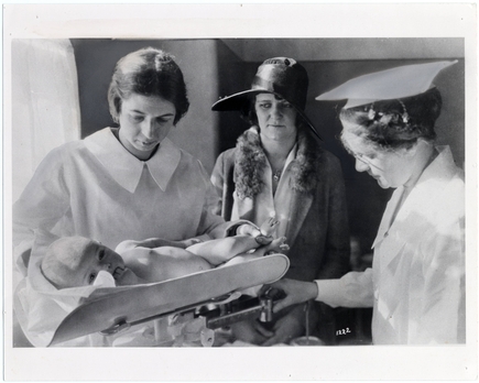 Dos enfermeras atienden a un bebé en una báscula mientras otra mujer con abrigo y sombrero observa; Fotografía en blanco y negro tomada en el hospital Doernbecher en la década de 1930.