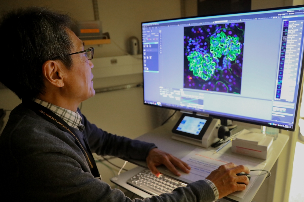 Nakai has short dark gray hair and is wearing a gray sweater over a dress shirt, working on a cell image on his computer.