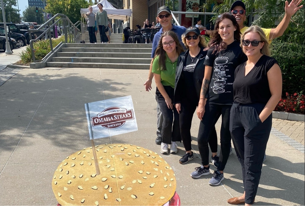 A group of people standing in front of a burger mural with Omaha Steaks flag