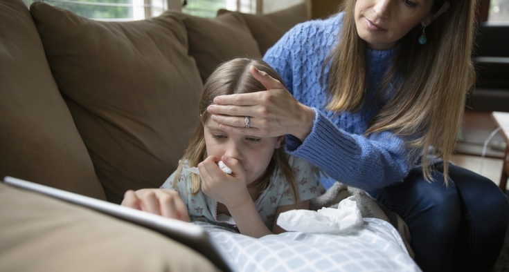 Mother checking forehead temperature of sick daughter laying on sofa