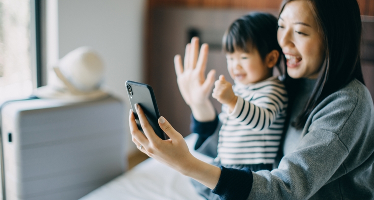 Young Asian mother and cute little daughter having video call on smartphone with family in hotel room while on vacation and smiling joyfully