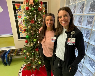Megan Areleth and Kristen Beaury, certified child life specialists at Novant Health Hemby Children's hospital, are smiling in front of a Christmas tree.