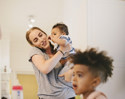 A mom holds her infant and talks on the phone while her toddler son sits in the foreground.