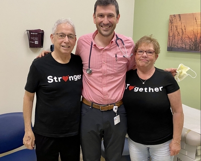 John and Jo Russo are wearing black shirts that say 'stronger together'. Dr. John Rommell is standing between then in a pink shirt with his stethoscope. All are smiling.