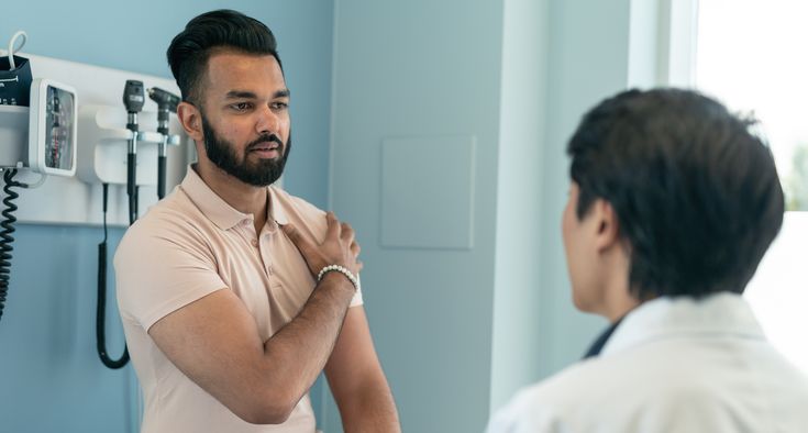 A man grips his shoulder with his hand while talking with a doctor in a white coat.