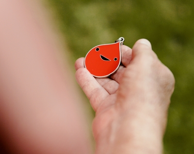 A hand holds a tiny keychain shaped like a drop of blood. It is red and smiling.