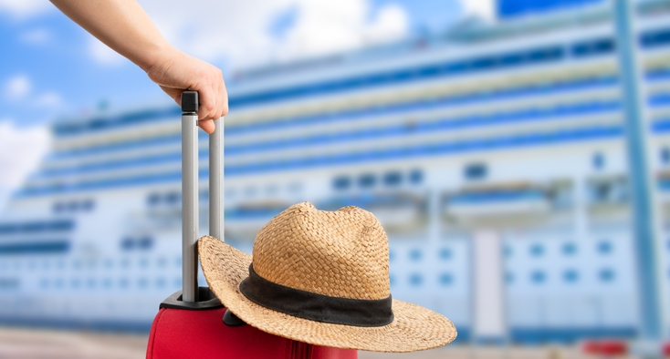A close up of a woman's hand holding the handle of a rolling suitcase and a straw sunhat. In the background, a large cruise ship is docked.
