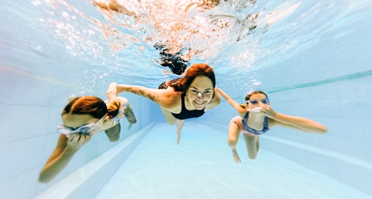 A woman swims underwater in a pool with two young girls in goggles swimming on either side of her.