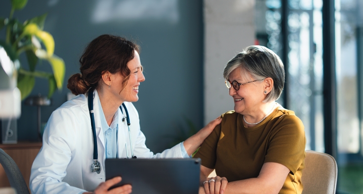 A senior woman and a doctor sit at a table facing each other and the doctor places her hand consolingly on the woman's shoulder.
