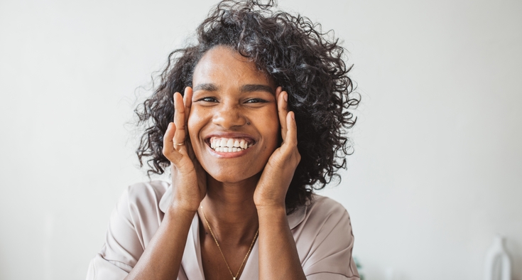 A woman with curly hair rests her fingertips on her temples while she smiles.