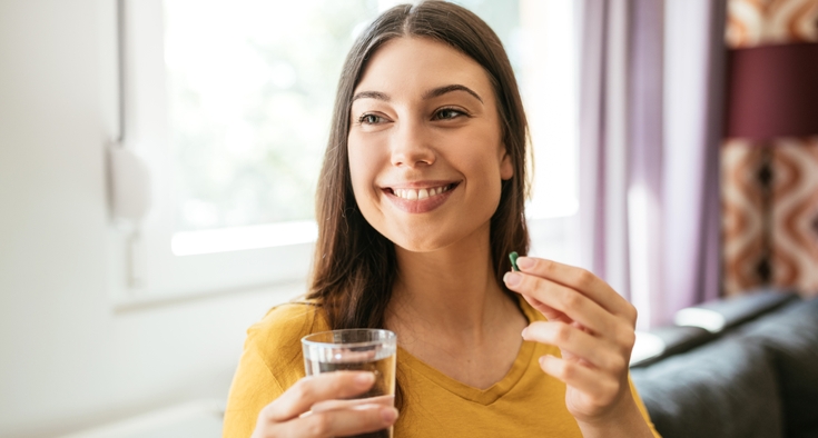 A woman in a yellow shirt smiles while holding a glass of water and a green supplement capsule.