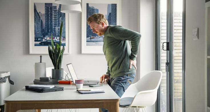 A man stands at his computer desk and clutches his lower back in discomfort.