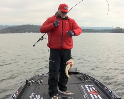 Hank Cherry stands on the hull of a fishing boat with a large fish hooked on the end of the line on his fishing rod.