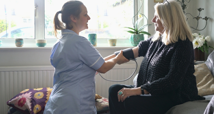 A nurse in blue scrubs uses a blood pressure cuff on a woman who is sitting on the couch in her home.